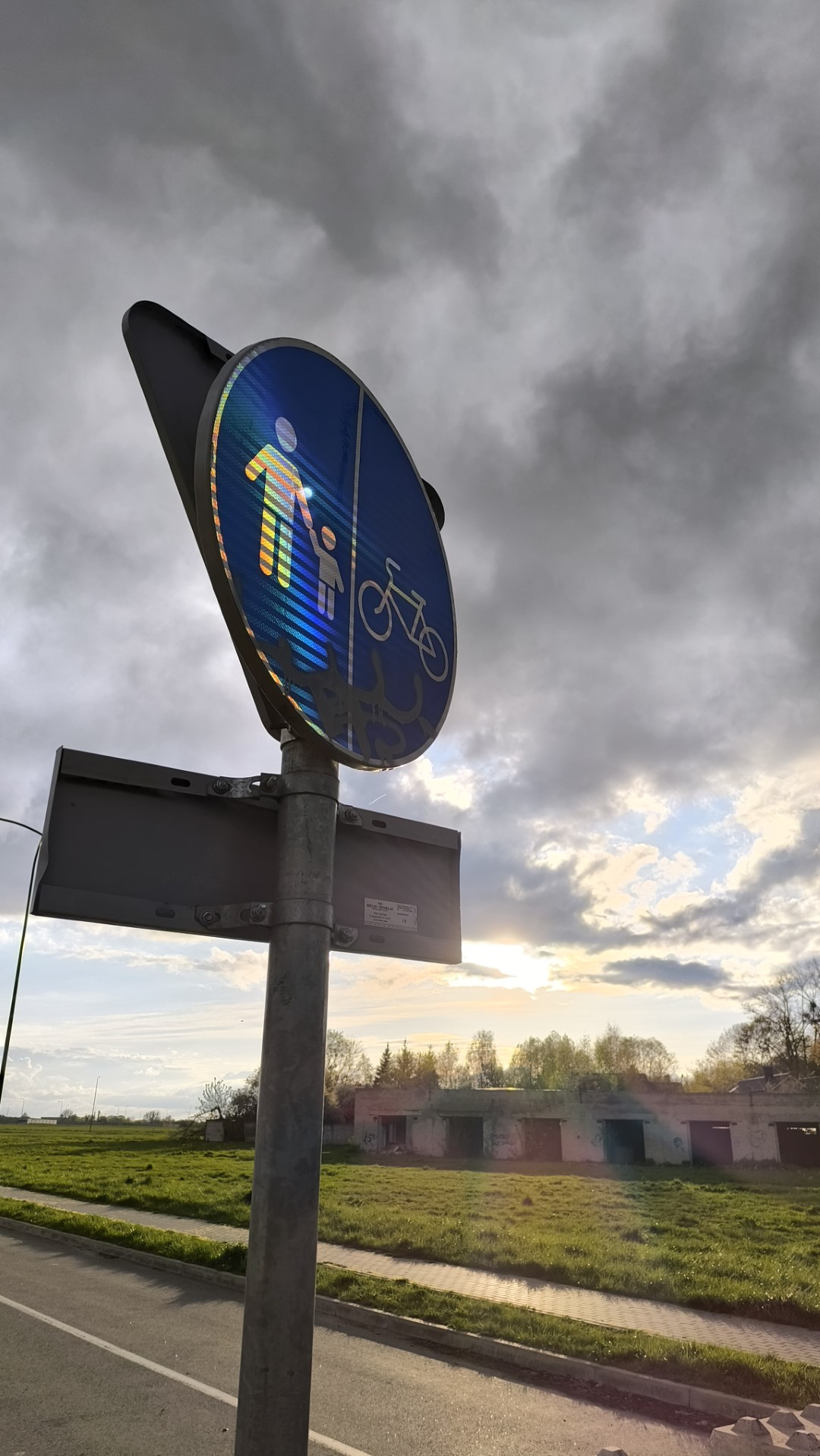 A photo of a street sign. The sun is setting in the background and is causing a rainbow to form on the street sign through reflections.